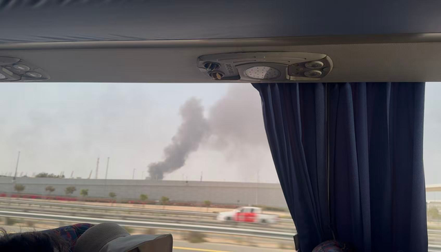 Thick black smoke rises into the sky behind a concrete wall along a highway, visible through a bus window with a curtain partially drawn.